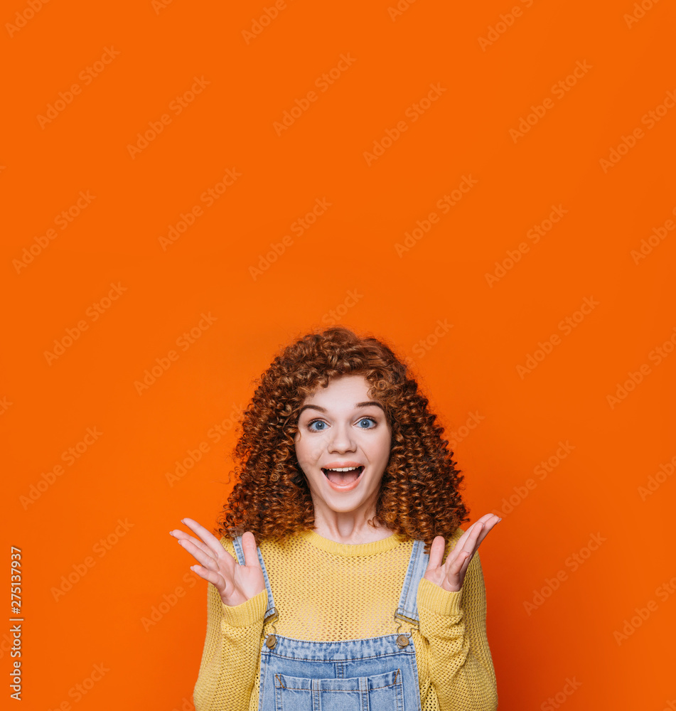 curly red-haired woman making shocked face and raised hands on orange background. Wow it amazing