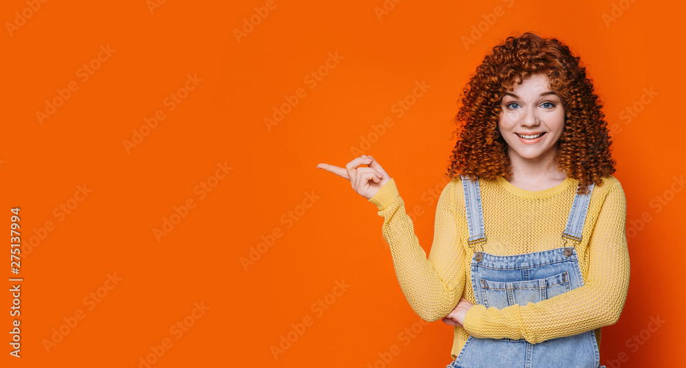 curly red-haired woman smiling and pointing hands into empty orange background.Hey you look this