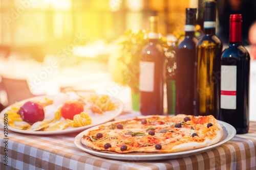 Italian pizza with tomatoes, mozzarella cheese, basil, black olives. In background, wine and highlight of light at sunset