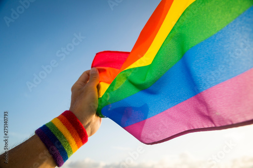 Hand with rainbow colors wristband waving gay pride flag waving backlit in the wind against a bright blue sky