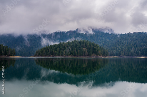 Montenegro, Black lake nature in durmitor national park landscape next to zabljak, green forested island mirroring on glassy calm lake water in mystic foggy atmosphere