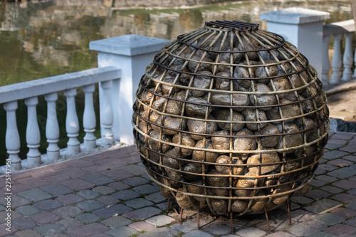 A ball of metal rods filled with stones against a white fence and pond.