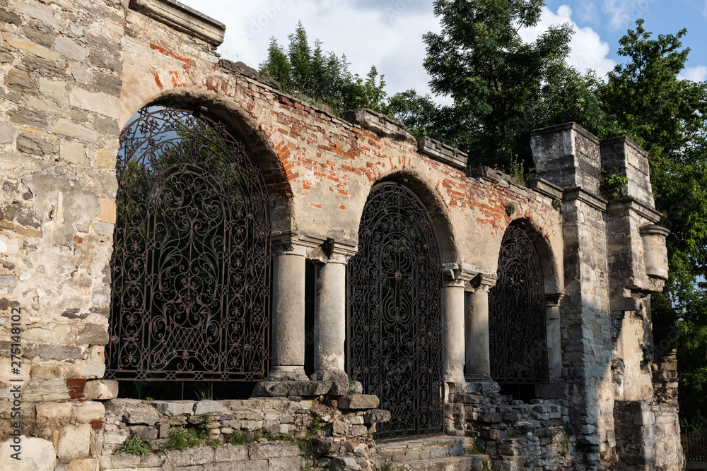 Part of a stone wall and a forged iron gate with columns in the background of greenery and sky. Ruins of the Armenian Church of St. Nicholas of the XV century, Kamianets-Podilskyi, Ukraine.