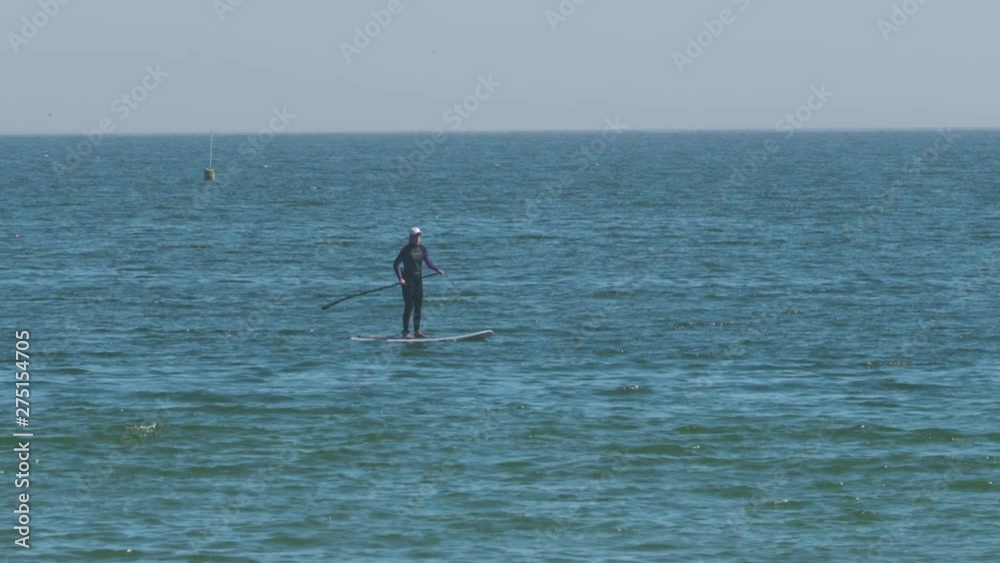 Handheld, medium wide shot of a person standing on a paddle board in the water. Handheld, medium wide shot of a person standing on a paddle board in the water.