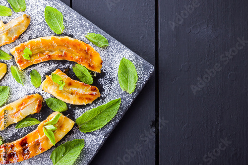 Halves of grilled bananas with powdered sugar on a black kitchen board and shale stone. Baked banana dessert with mint on black background table