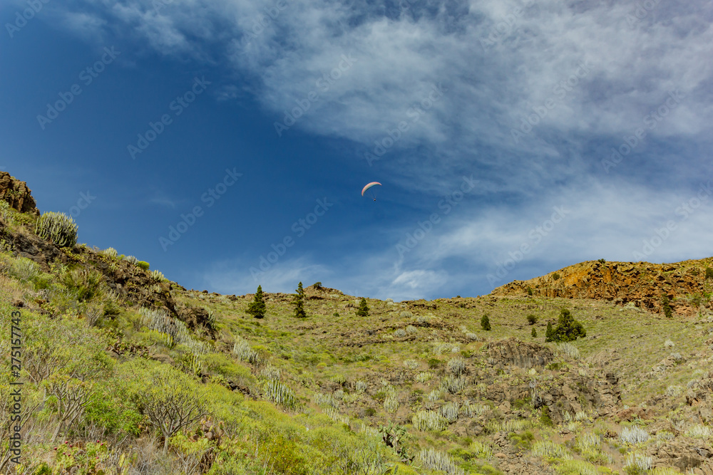 paraglider flying over the mountains peaks against the bright blue sky. Sunny day. Clear blue sky and some clouds above the mountains. Rocky tracking road in dry mountain area. Tenerife