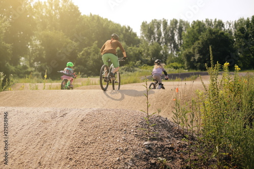 mother and two daughters riding bike together on a bicycle dirt track in bright summer light - family lifestyle outdoor activity concept - focus on hill tip in foreground - rider blanked out blurry