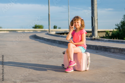 Little girl sitting on a pink suitcase near the road, free space.