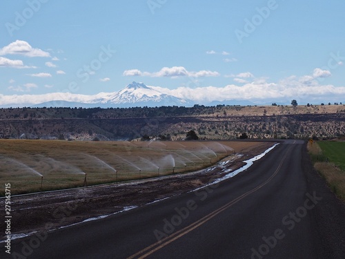 A rural road running through farmland being irrigated in Central Oregon with snow covered Mt. Jefferson in the background on a sunny summer day.