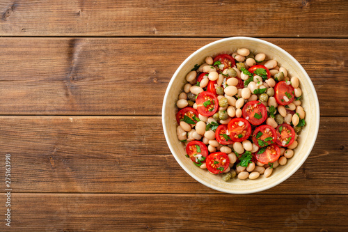 White bean salad with tomatoes, capers, garlic and parsley in bowl on wooden table. Top view. Copy space.