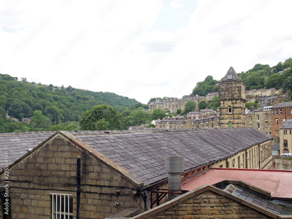 a view of the steep hillside streets in hebden bridge between summer trees with the tower of the historic nutclough mill building at the front