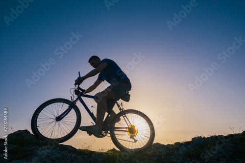 Fototapeta Naklejka Na Ścianę i Meble -  Silhouette of a fit male mountain biker riding his bike uphill on rocky harsh terrain on a sunset.