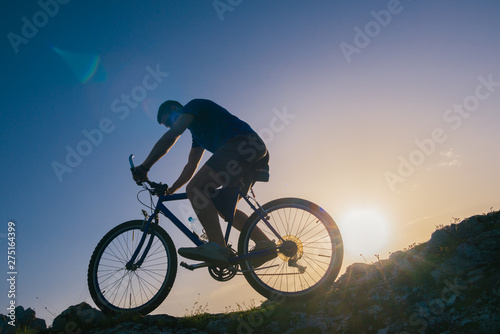 Fototapeta Naklejka Na Ścianę i Meble -  Silhouette of a fit male mountain biker riding his bike uphill on rocky harsh terrain on a sunset.