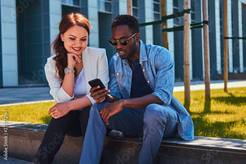 Happy smiling multicultural couple are wathing something on mobile phone while sitting on the grass near building. photo