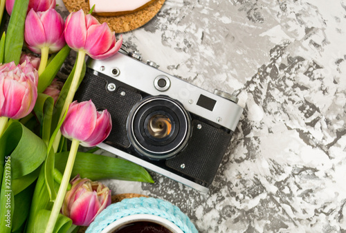 The old camera is lying on a concrete table, next to a cup with hot tea, next to it are a pink bouquet of tulip flowers. Concept breakfast, instagram, travel.