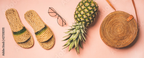 Variety of summer apparel items. Flat-lay of summer flip flops, sunglasses, wicker shoulder bag and pineapple over pastel pink background, top view, wide composition. Summer fashion garment concept