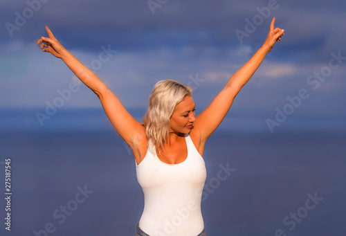 young attractive and beautiful 30s blond hair woman in casual top and denim shorts spreading arms happy and cheerful posing isolated on sea blue and sky