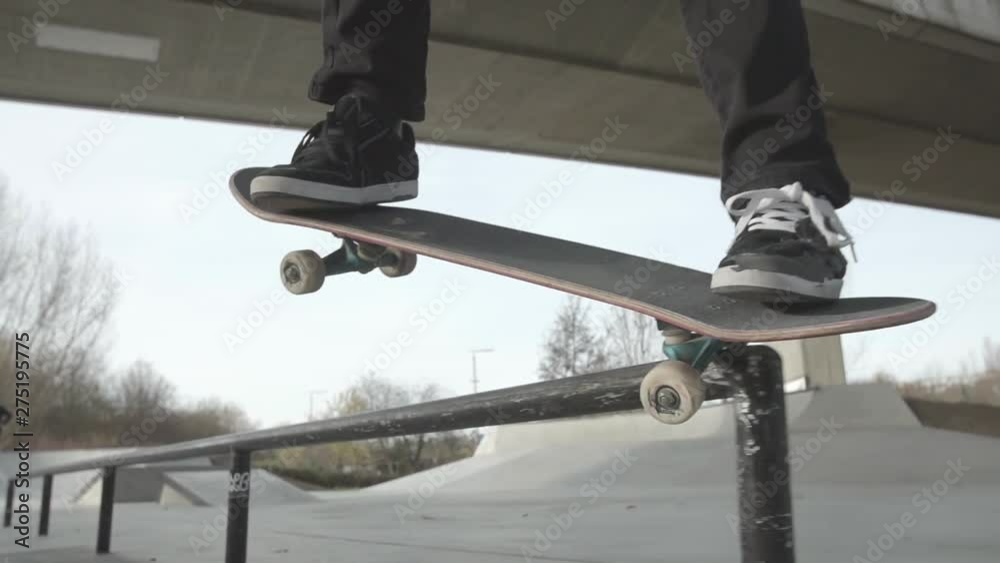 A skater does a backside crooked grind on a flat rail at a concrete skatepark. The shot is in slow-motion, outdoor, blue sky.