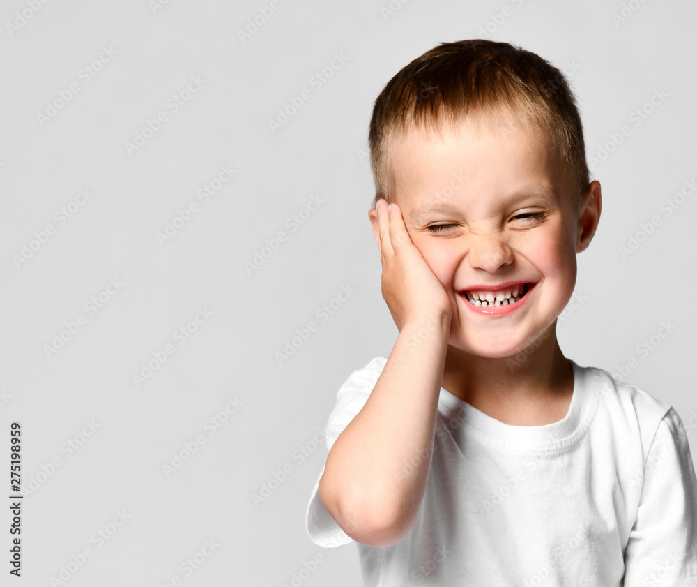 emotional portrait of a little boy in a white t-shirt. The boy looking at the camera, put his palm on his cheek, hesitates and laughs at the one who has been seen. emotional portrait of a little boy in a white t-shirt. The boy looking at the camera, put his palm on his cheek, hesitates and laughs at the one who has been seen.