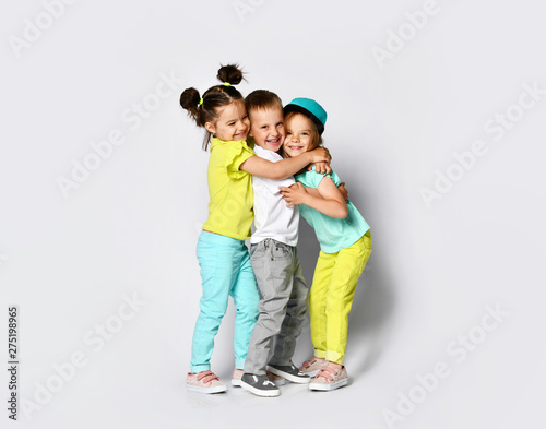 Studio portrait of children on a light background: full body shot of three children in bright clothes, two girls and one boy. Triplets, brother and sisters. hugging on camera. Family ties, friendship