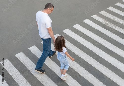 A man and a small child on a zebra crossing trespassing by crossing the street. In the summer on the street kid girl with her father in fashion clothes cross the road.  From top view