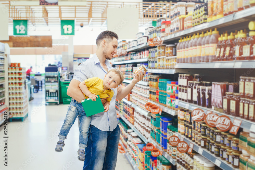 Caucasian father dad in grocery store shopping buying food and holding carrying baby son under arm. Man parent with toddler kid choosing meal for dinner lunch. Difficult parenting authentic moment.