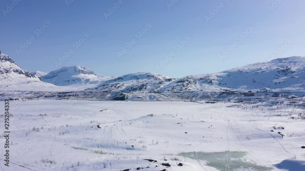Aerial view of small town by frozen lake in snowy mountain landscape. Blue sky. MOVING FORWARD AND DOWN.