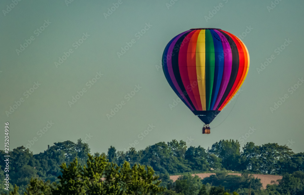 Obraz premium Rainbow colored hot-air balloon floats over the hiltop forest in early morning spring with cloudless sky