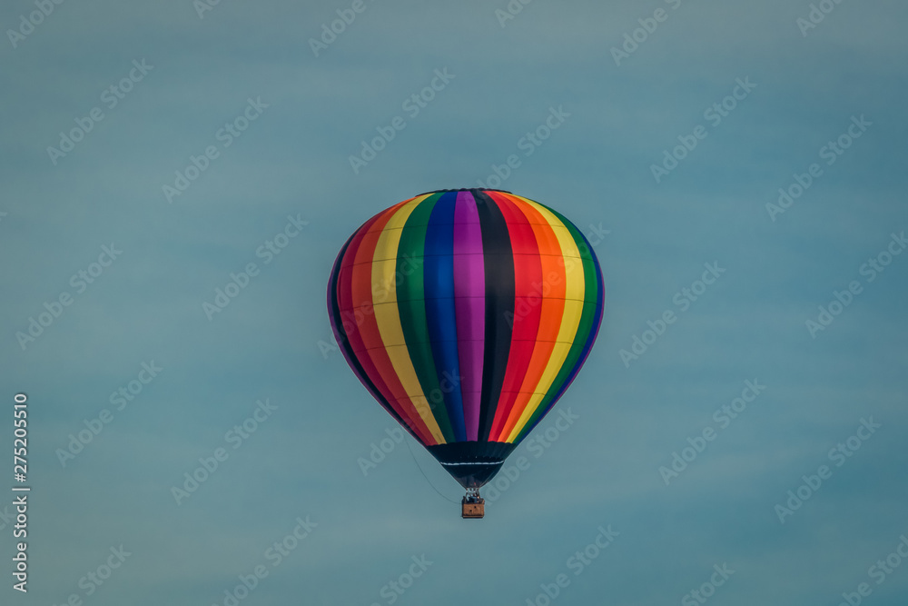 Fototapeta premium Rainbow colored hot-air balloon floats over the hiltop forest in early morning spring with cloudless sky