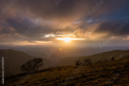 Moody sunset on Monte Cucco (Umbria, Italy), with tree in the foreground and sun filtering through clouds