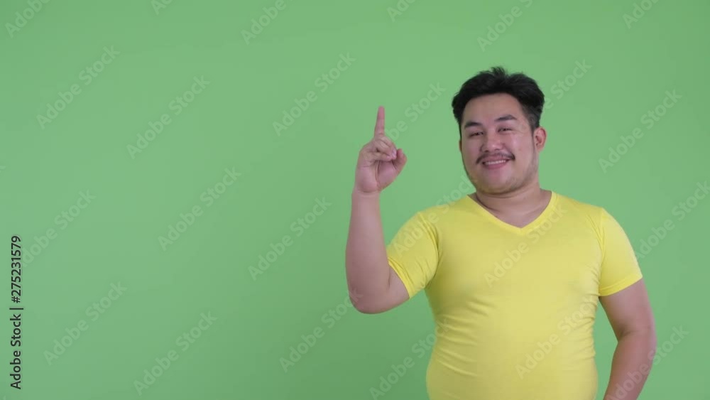 Happy young overweight Asian man talking while pointing up Happy young overweight Asian man talking while pointing up