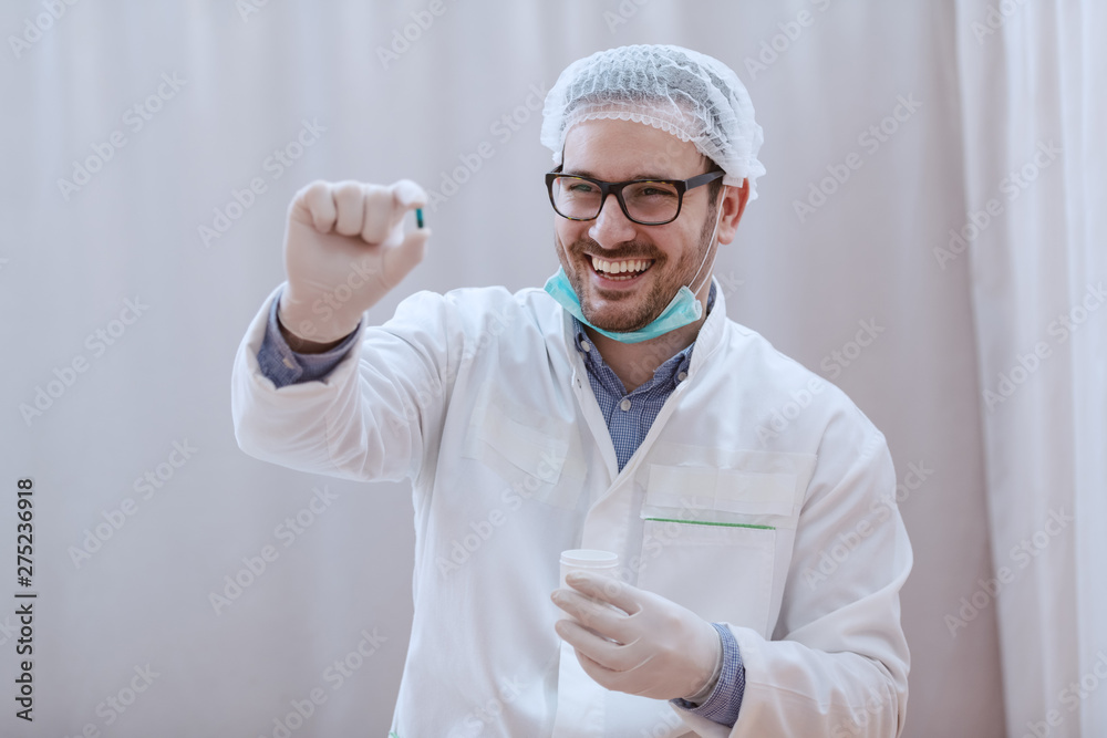 Young smiling Caucasian doctor in white uniform, mask and hairnet holding capsule. Selective focus on hand.