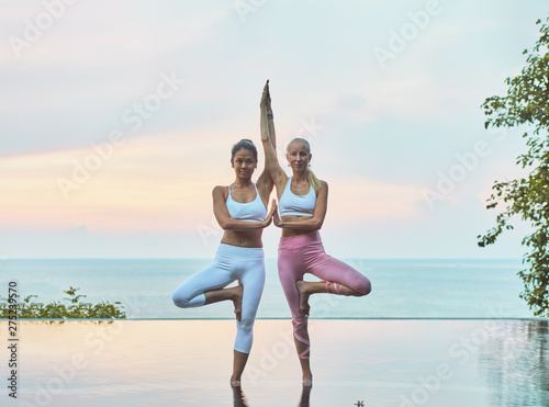  Two young women doing yoga in a pool by the sea at sunrise 