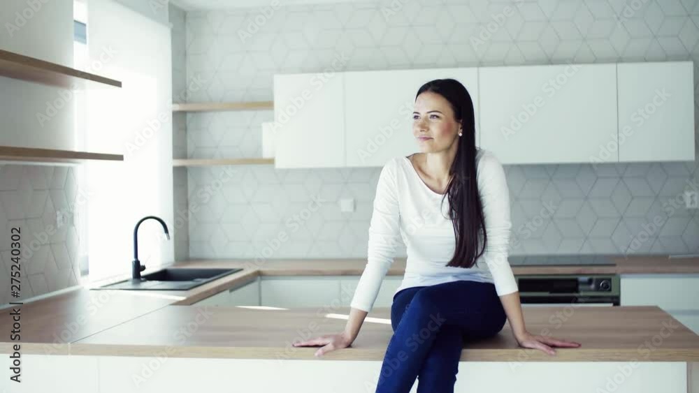 A cheerful young woman sitting on kitchen counter in new home, resting. A cheerful young woman sitting on kitchen counter in new home, resting.
