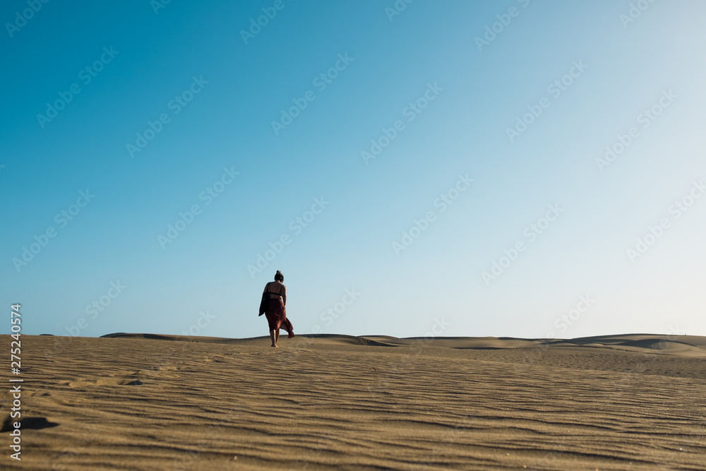 young confident woman walking alone barefoot on path in desert sand among dunes on a hot sunny day with clear blue sky
