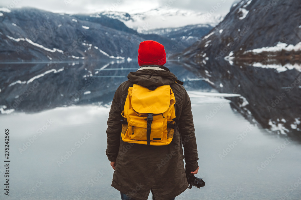 Traveler man with a yellow backpack wearing a red hat standing on the background of mountains and lake enjoying landscape. Travel lifestyle concept. Shoot from the back