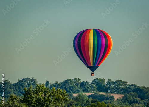 Rainbow colored hot-air balloon floats over the hiltop forest in early morning spring with cloudless sky © rabbitti
