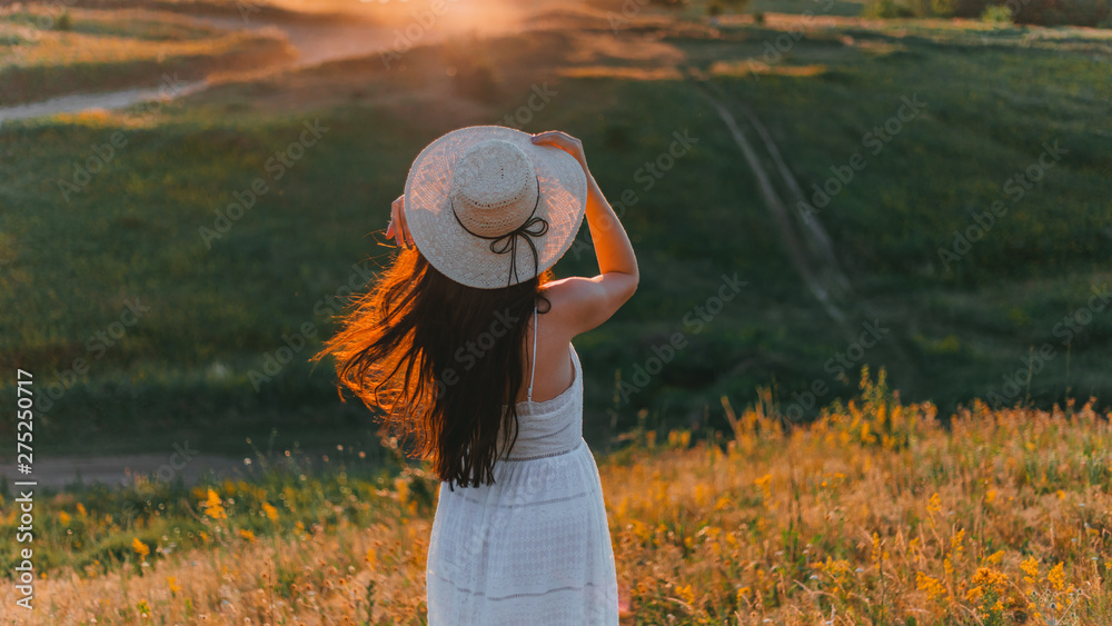 A young girl with black hair in a hat and white dress stands with his back in the meadow and looks at the sunset. Concept - Freedom and Unity with Nature