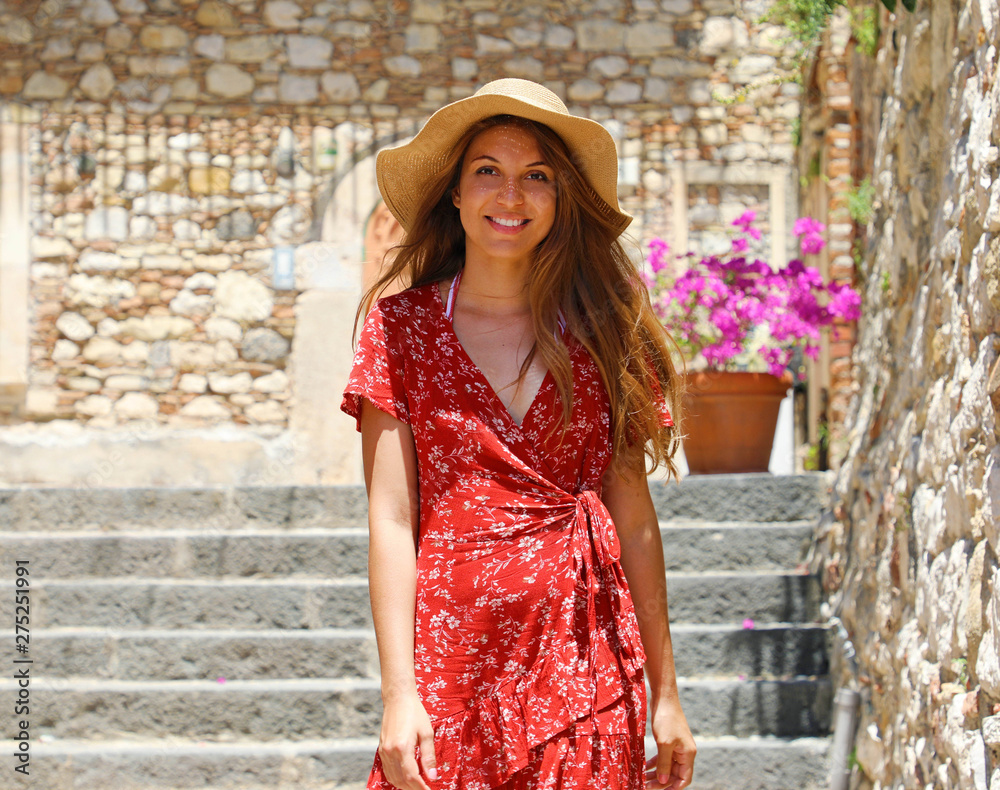 Beautiful tourist girl with hat and red dress walking in cozy italian street in Taormina, Sicily, Italy