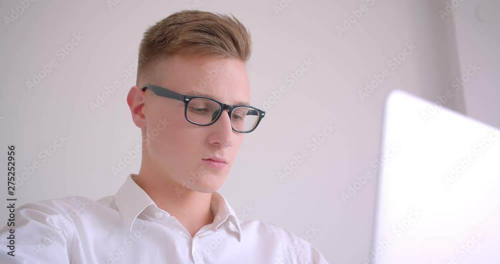 Closeup portrait of young handsome caucasian businessman in glasses using the laptop looking at camera smiling confidently sitting in the armchair indoors in a white room Closeup portrait of young handsome caucasian businessman in glasses using the laptop looking at camera smiling confidently sitting in the armchair indoors in a white room