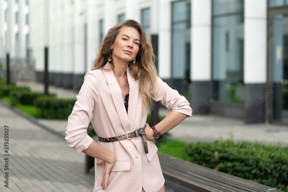 Fototapeta premium Beautiful girl posing on the street in a coral jacket and a pleated skirt. Long flowing hair. Horizontal portrait.