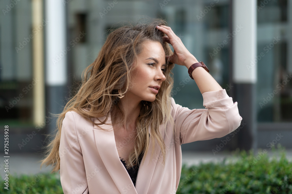 close-up portrait of a beautiful natural girl with flowing hair in a coral jacket. On the street against the glass showcases of the business center. Summer day.