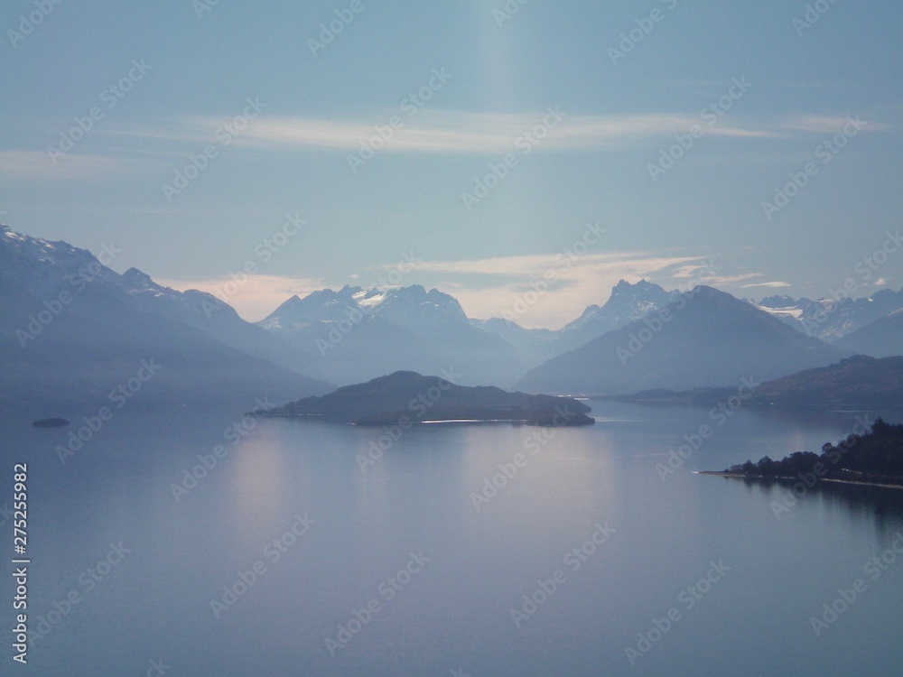 A silver grey lake is surrounded by mountains. Some of the mountains have snow. A tree-covered island is in the lake. The sky is blue, with some clouds.