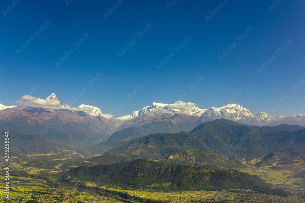 city in a bright green mountain valley on the background of wooded hills and snowy peaks of Annapurna with white clouds under a clear blue sky