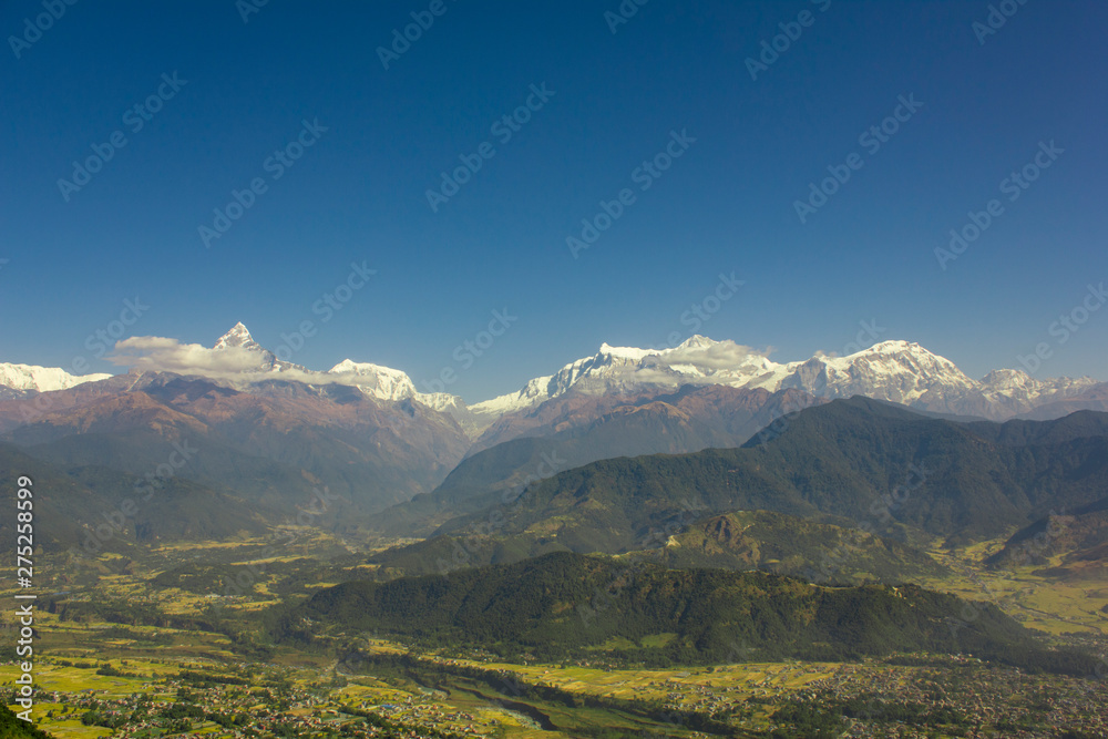 village with a river in a green mountain valley against the wooded slopes and snowy peaks of Annapurn in white clouds under a blue sky