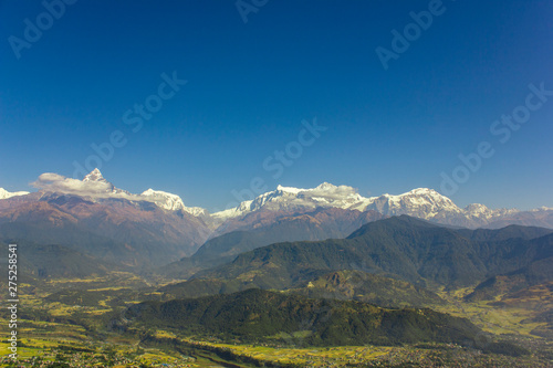 city in a bright green mountain valley on the background of wooded hills and snowy peaks of Annapurna with white clouds under a clear blue sky