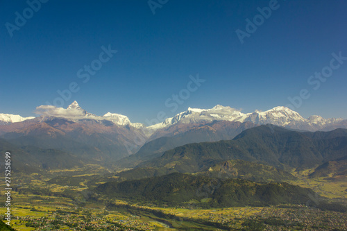 village with a river in a green mountain valley against the wooded slopes and snowy peaks of Annapurn in white clouds under a blue sky © Pavel