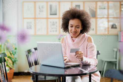 Beautiful mixed race woman in pink turtleneck sweater sitting in pastry shop and using laptop for on line shopping.