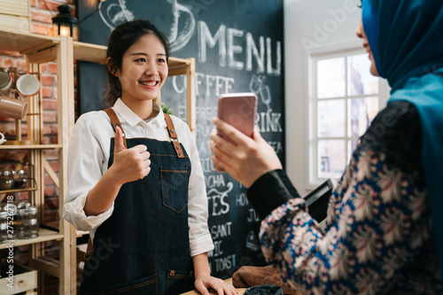 muslim woman holding mobile phone showing screen with pay bill at counter coffee shop. Digital lifestyle online payment concept. young girl waitress staff ok good thumb up hand sign gesture in cafe