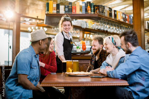 Portrait of smiling young diverse friends in indoors restaurant and smiling waitress with drinks and beer.